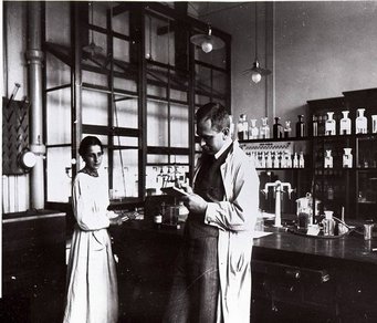 Lise Meitner und Otto Hahn im Berliner Labor in 1913. Bild: Archiv der MPG Berlin-Dahlem Zwei Personen in weißen Kitteln in einem historischen Labor mit Glasflaschen und Geräten.