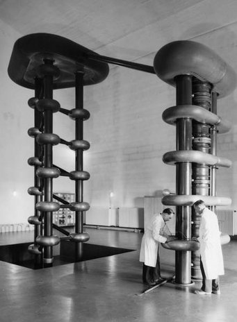 Die Hochspannungskaskade mit Ionenrohr wurde am MPI für Chemie bis 1980 als Teilchenbeschleunigergenutzt. Bild: MPI für Chemie Zwei Forscher in weißen Kitteln stehen vor einer großen Hochspannungsanlage mit mehreren isolierenden Ringen und einer Querstange in einem Laborraum.