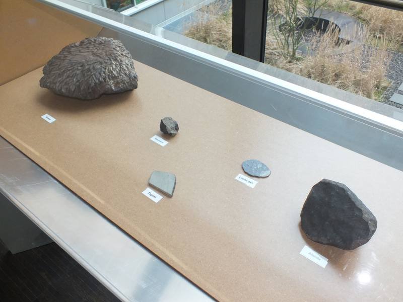 Eine Museumsvitrine zeigt fünf Meteoriten unterschiedlicher Größe und Farbe, jede mit einem Etikett zur Identifikation.
