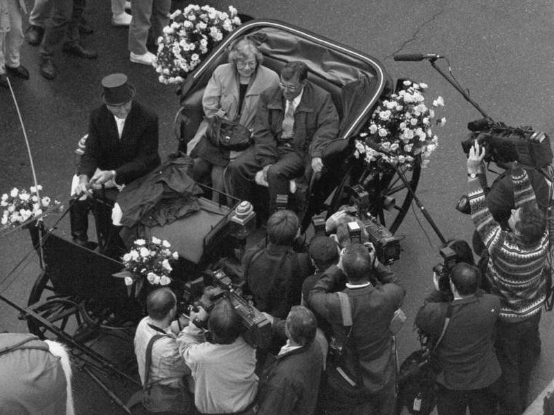 Schwarz-weiß Foto einer Pferdekutsche in der 2 Personen hinten und eine Person auf dem Kutschbock sitzt. Außen herum stehen mehrere Journalisten mit Foto- und Videokameras.