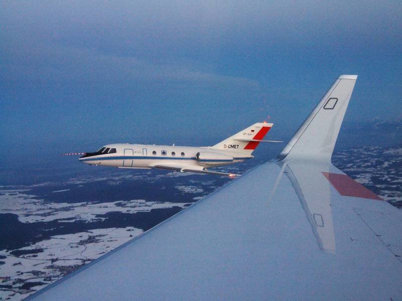 Sicht auf ein zweites Flugzeug neben dem Flügel eines Flugzeugs über einer winterlichen, verschneiten Landschaft bei Abenddämmerung.