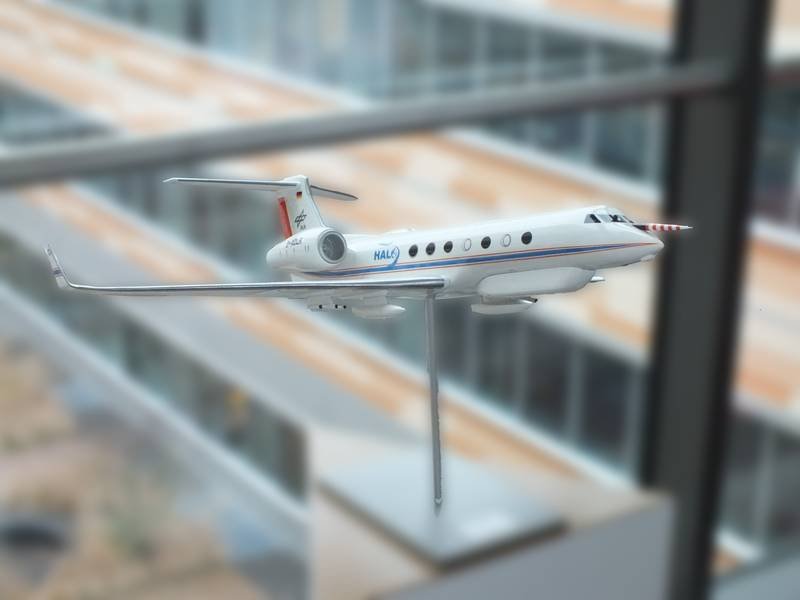 Detailliertes Modell eines weißen Flugzeugs mit roten und blauen Streifen, montiert auf einem Ständer vor einem unscharfen Hintergrund.