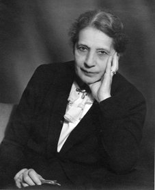 Lise Meitner (1878-1968). Quelle: MPG Archiv Berlin-Dahlem Schwarz-weißes Porträt einer Frau in einem Anzug, die einen Stift hält und den Kopf auf eine Hand stützt.