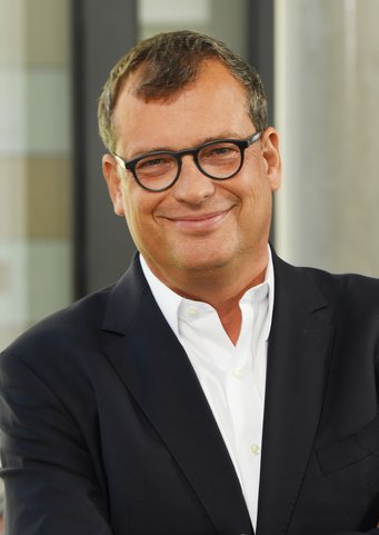 Gerald H. Haug Man dressed in a dark blazer over a white button-up shirt, standing with crossed arms in front of a blurred background.