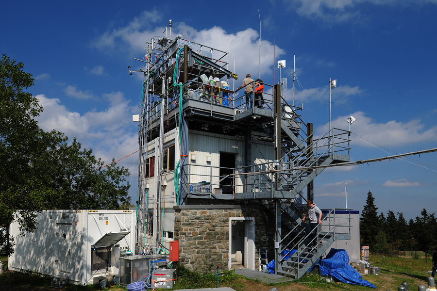 Mehrstöckiger Turm mit Wettermessgeräten, Antennen und Kabeln, auf dem mehrere Personen auf der Treppe und Plattform technische Arbeiten durchführen.