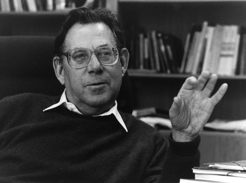 Man sitting in an office chair with bookshelves in the background, raising one hand.
