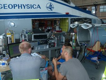 Technicians analyze equipment under Geophysica aircraft in a hangar setting.