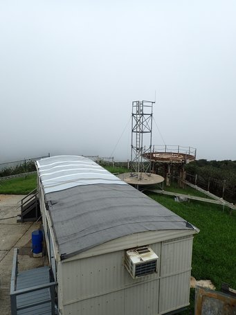 Die Messstation „Pico del Este“ vor (o.) und nach (u.) dem verheerenden Hurrikan „Maria“, der mit 250 Kilometern pro Stunde Mitte September über die Insel fegte. Weißes Gebäude mit gewölbtem Dach, Klimaanlage an der Seite, neben einem hohen Metallturm im Nebel, umgeben von grüner Landschaft.