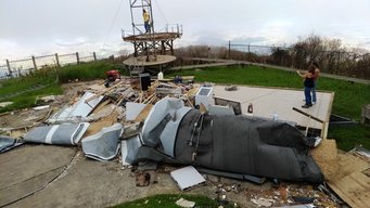 Messstation „Pico del Este“ nach Hurrikan "Maria". Trümmerteile neben einem Turm auf einer grünen Fläche, Person steht daneben.