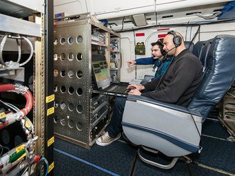 Überwachen die Messungen des Aerosolteilchenmassenspektrometers ERICA: Andreas Hünig (l.) und Antonis Dragoneas. Eine Person mit Kopfhörern sitzt in einem Flugzeugsitz und bedient einen Laptop, umgeben von technischen Geräten und Kabeln in einer Flugzeugkabine.