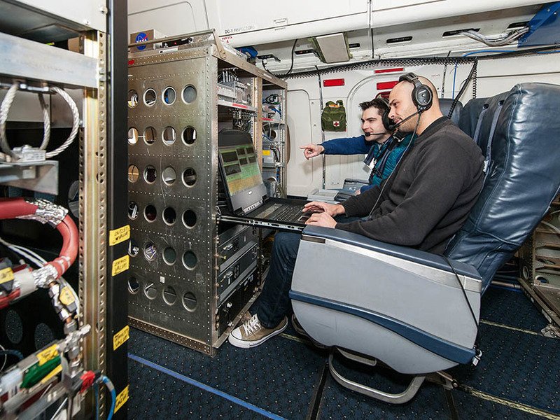 Andreas Hünig (left) and Antonis Dragonea (right) in aircraft seats using a laptop, wearing headphones, with equipment racks visible.