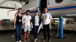 Von links: Bruna Holanda, Ovid Krüger, Katharina Kaiser und Johannes Schneider posieren vor einem Flugzeug, das in einer großen Halle steht. Im Hintergrund befindet sich technische Ausrüstung, einschließlich Druckflaschen und Schläuchen.