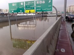 Urban highway scene with significant flooding, causing road signs for Al Ruwalis, Madinah, and Al Balad to be partially submerged, with vehicles parked on the side.