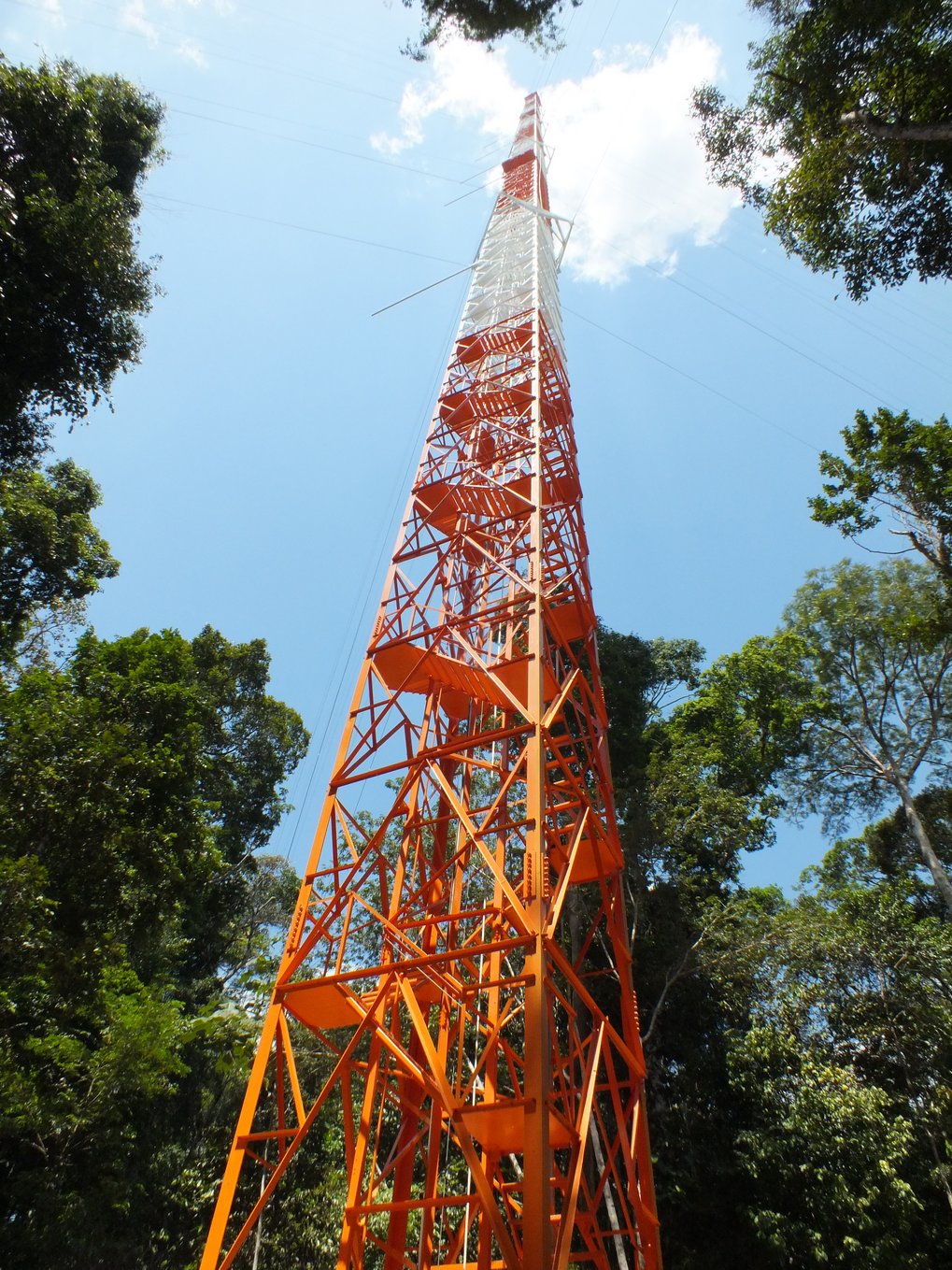 Orange and white metal communication tower with platforms, set against a blue sky and green trees.