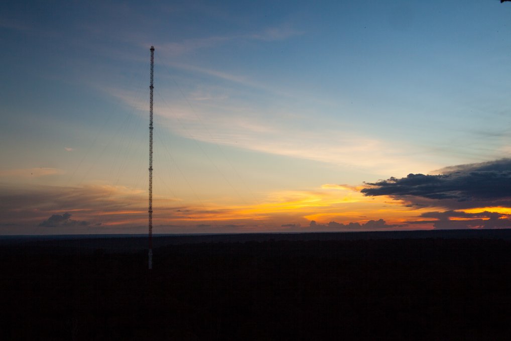 A slender mesh metall tower stands alone on the horizon with a vibrant sunset featuring orange, yellow, and blue hues.
