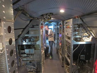 Mira Pöhlker aboard the research aircraft HALO. Photo: Ulrich Pöschl Interior of an aircraft featuring multiple racks filled with electronic equipment and wiring, with Mira Pöhlker inspecting a device.