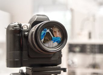 Quelle: MPIC/MPIP Kameraobjektiv spiegelt Forscherin mit blauen Handschuhen wider.