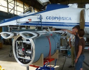Einbau des Aerosolpartikel-Massenspektrometers ERICA in das Forschungsflugzeug M-55 Geophysica. (Foto: S. Borrmann, IPA, MPIC) Ein Flugzeug mit der Aufschrift „Geophysica“ im Hangar, ausgestattet mit einem großen wissenschaftlichen Instrument, während drei Personen das Gerät betrachten und diskutieren.