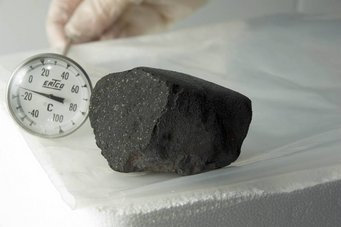 Der Tagish Lake-Meteorit enthält große Mengen an kohlenstoffhaltigem Material. Foto: Michael Holly, Creative Services, University of Alberta Ein dunkler, poröser Stein liegt neben einem analogen Thermometer, das Temperaturen von -50°C bis +100°C auf einer weißen Fläche misst. Eine behandschuhte Hand ist im Hintergrund sichtbar.