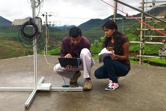 Sachin S Gunthe (l.) und Akila Muthalagu während der Messungen in Indien. Foto: Gerhard Lammel Zwei Personen knien auf einer Betonfläche und bedienen einen Laptop, umgeben von grünen Hügeln.