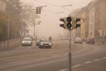 Source:Martin Vorel/Wikimedia Commons A street with a green traffic light in foggy conditions, featuring several cars, tram tracks, and buildings lining both sides of the road.