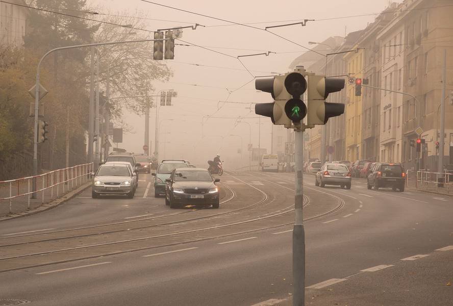 Eine Straße mit einer grünen Ampel bei Nebel, auf der mehrere Autos fahren und an beiden Seiten der Straße Straßenbahnschienen und Gebäude zu sehen sind.