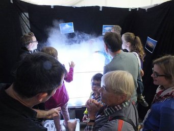 Die Eigenschaften von Wolken werden für jedermann begreifbar gemacht mithilfe einer Wolkenkammer am Tag der Deutschen Einheit in Mainz. Foto: Benner A group of individuals stands around a misty light display, possibly examining a scientific experiment.