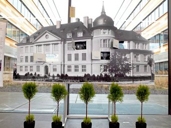 Model of the old KWI for Chemistry building in the entrance area of the new MPIc building. Photo: S. Benner Historical black-and-white image of a large building mounted on a glass panel inside a modern building, with five small potted trees aligned in front.
