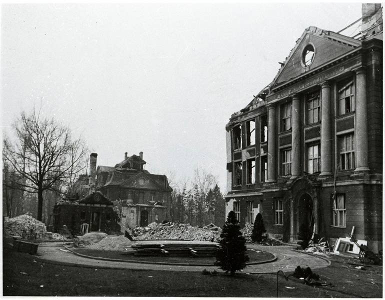 Damaged classical buildings with rubble scattered around and leafless trees in the background.