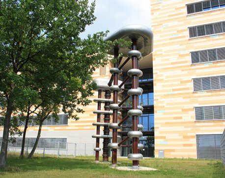 Outdoor scientific apparatus featuring tall vertical rods and metallic rings, positioned on a lawn next to a modern office building with trees nearby.