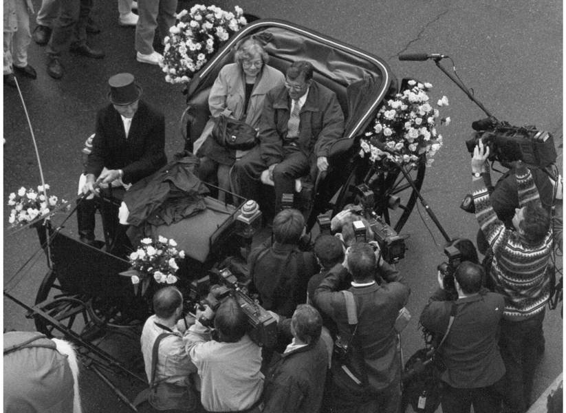 A black-and-white photo showing a carriage from above. Two people are sitting in the back of the carriage, and one person is sitting on the driver's seat in the front. The carriage is surrounded by journalists with cameras.