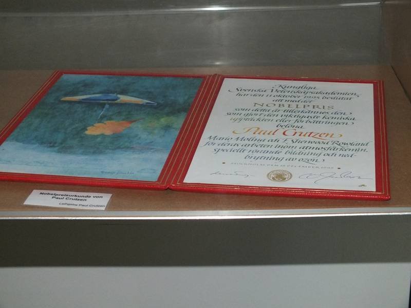 Red-framed certificate and a painting are exhibited side by side in a glass case, highlighting an official recognition.