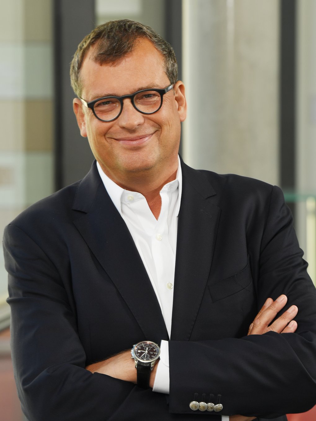 Man in a dark suit with arms crossed, wearing a white shirt and a wristwatch.