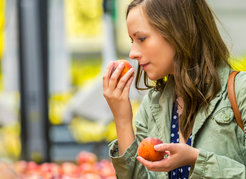 Woman in a green jacket holding apples at a grocery store, surrounded by fruit. She is holding one of the apples close to her nose.