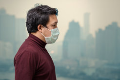 A man in a dark red sweater and face mask stands in front of a hazy cityscape, with tall buildings partially visible in the background.