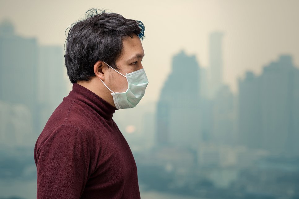 Eine Person mit dunklem Haar und Schutzmaske blickt zur Seite, während im Hintergrund eine verschwommene urbane Skyline zu sehen ist.