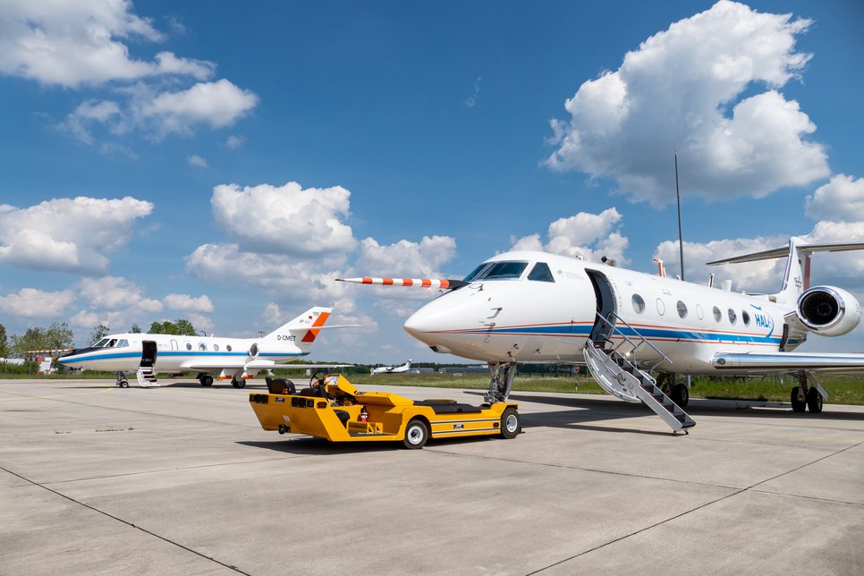Auf einem Flugfeld befinden sich zwei Flugzeuge. Im Vordergrund zieht ein gelbes Bodenfahrzeug ein Flugzeug. Der Himmel ist teils bewölkt.
