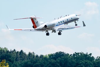 Start des Forschungsflugzeugs HALO im Rahmen der Mission BLUESKY vom DLR-Standort Oberpfaffenhofen. Quelle: DLR Ein zweistrahliges Flugzeug mit roten und blauen Streifen hebt über einem Wald ab, während es in den Himmel steigt.