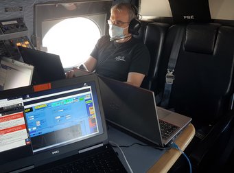 Der erste Flug des Forschungsflugzeugs HALO im Rahmen der Mission BLUESKY. Thomas Klimach überwacht per Computer die Messinstrumente. Quelle: MPIC Zwei geöffnete Laptops auf einem Tisch in einem Flugzeug mit Kontrollpanel. Mann mit Maske und Kopfhörern bedient einen der Laptops.