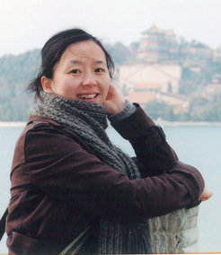 Forscherin Yafang Cheng in brauner Jacke und grauem Schal steht vor einem See mit einem Tempel im Hintergrund.