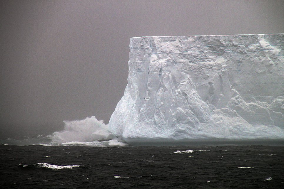 Massive iceberg with jagged edges amidst stormy seas and crashing waves.
