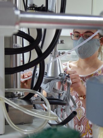 A fabric mask is tested for its filtration efficiencies. Source: Frank Drewnick Detailed view of laboratory equipment featuring interconnected tubes and metal structures, with a female researcher adjusting components.