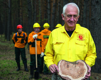Feuerökologe Prof. Dr. Dr. h.c. mult. Johann Georg Goldammer. Johann Goldammer in gelber Uniform mit „Global Fire Monitoring Center“-Logo hält Holzscheibe, während vier Personen in Schutzkleidung, darunter Helme, im Wald stehen.