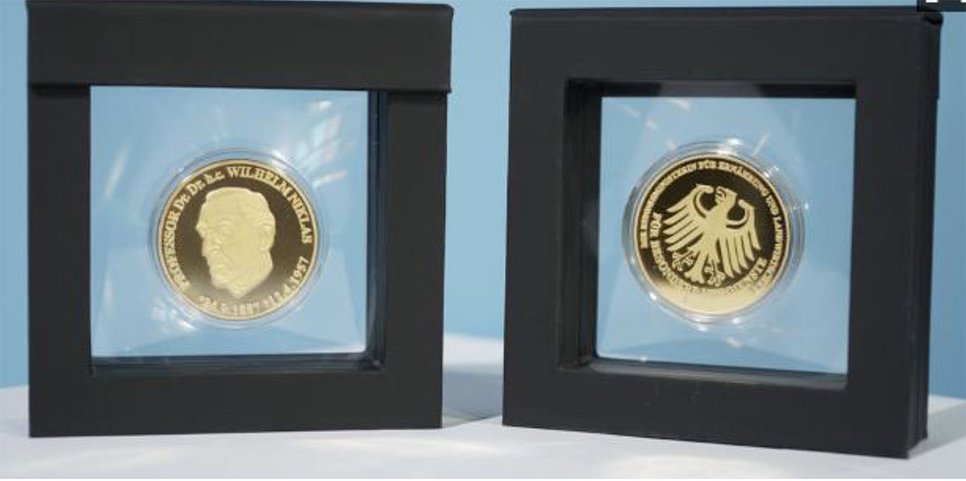Zwei goldene Münzen in schwarzen Rahmen, links mit einem Porträt und Text, rechts mit einem Adler und Schriftzug. Beide Münzen sind in transparenten Hüllen präsentiert.