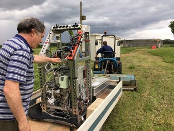 Forscher des MPI für Chemie und der Universität von Pisa installieren das PTR-MS für die Feldmessungen. Eine Person bedient ein komplexes Gerät auf einem Anhänger, während ein Traktor im Hintergrund auf einem Feld steht, umgeben von bewölktem Himmel.