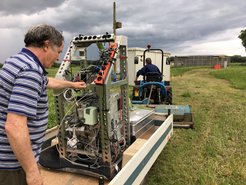 Eine Person bedient ein komplexes Gerät auf einem Anhänger, während ein Traktor im Hintergrund auf einem Feld steht, umgeben von bewölktem Himmel.