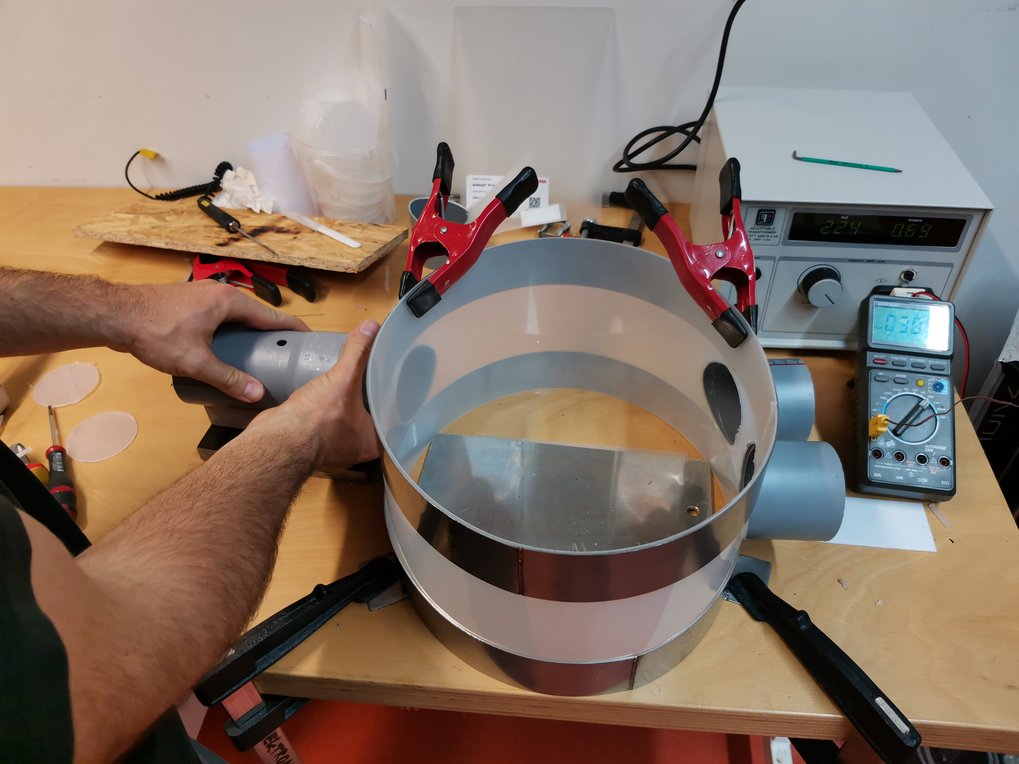 Ein Mann arbeitet an einem zylindrischen Gerät auf einer Werkbank, umgeben von Klemmen, Werkzeugen und einem digitalen Multimeter. Ein Netzgerät zeigt Messwerte an.