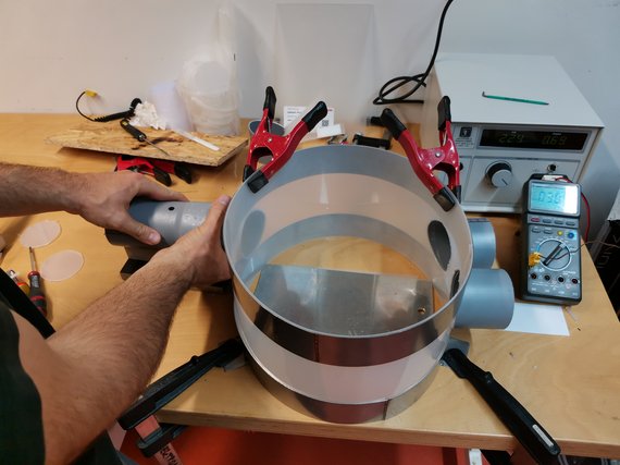 Ein Mann arbeitet an einem zylindrischen Gerät auf einer Werkbank, umgeben von Klemmen, Werkzeugen und einem digitalen Multimeter. Ein Netzgerät zeigt Messwerte an.