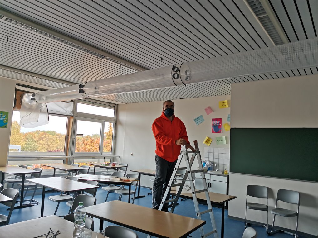 Klassenzimmer mit Tischen, Stühlen und einer Leiter unter einer Decke mit Belüftung.