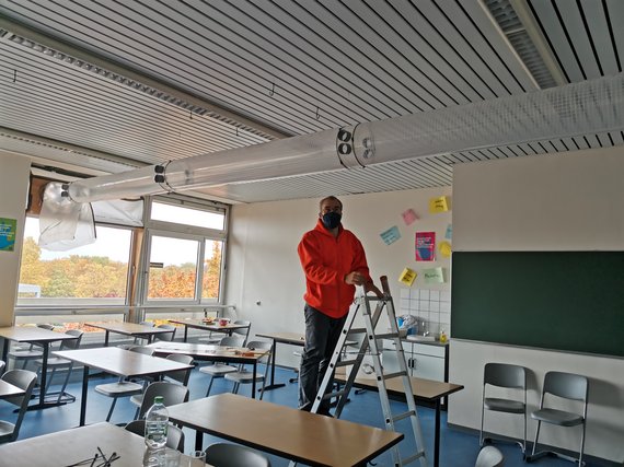 Klassenzimmer mit Tischen, Stühlen und einer Leiter unter einer Decke mit Belüftung.